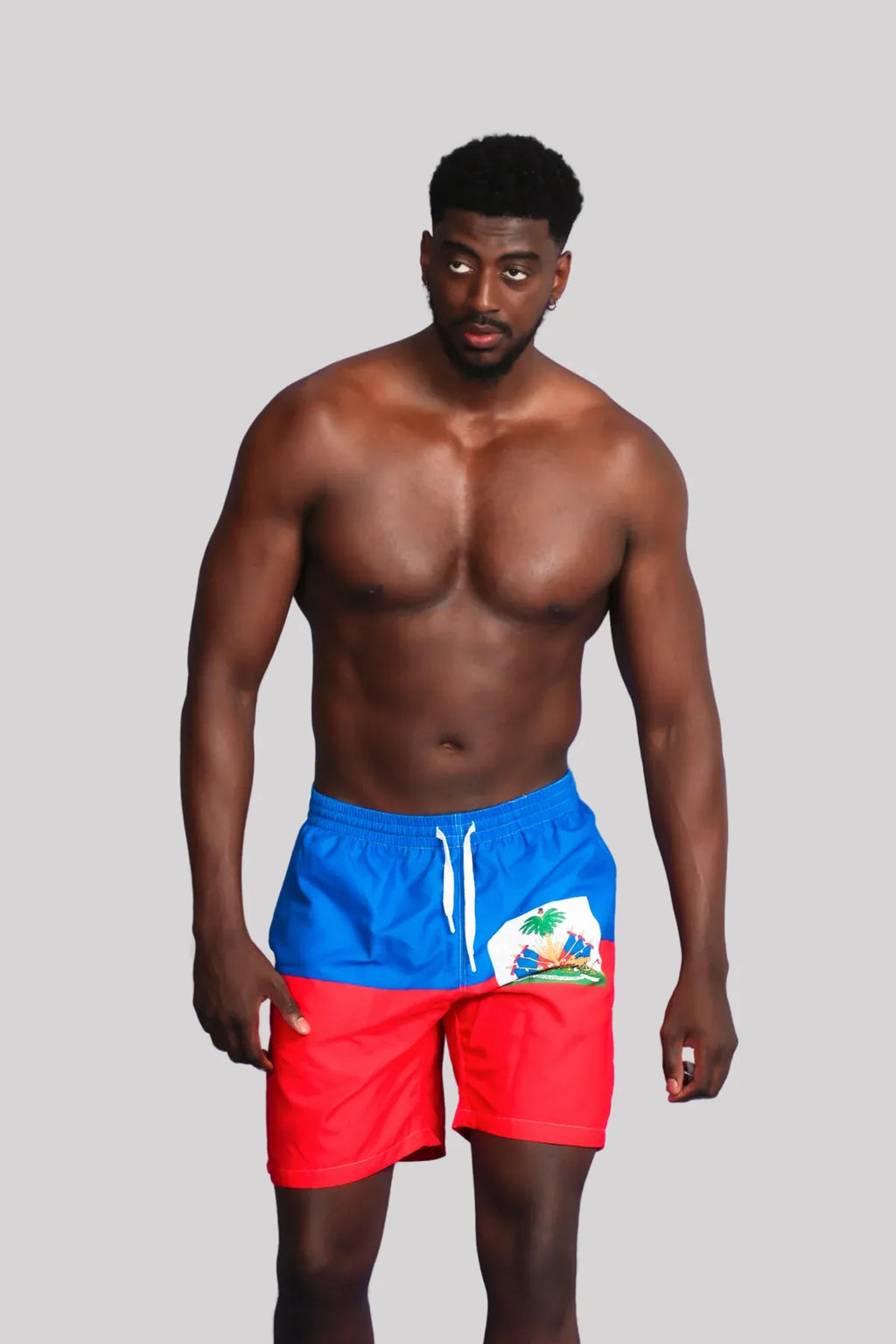 Model Jimseuh wearing Haiti flag swim shorts in red and blue with coat of arms on front — bold, culturally-inspired menswear by Travaly.