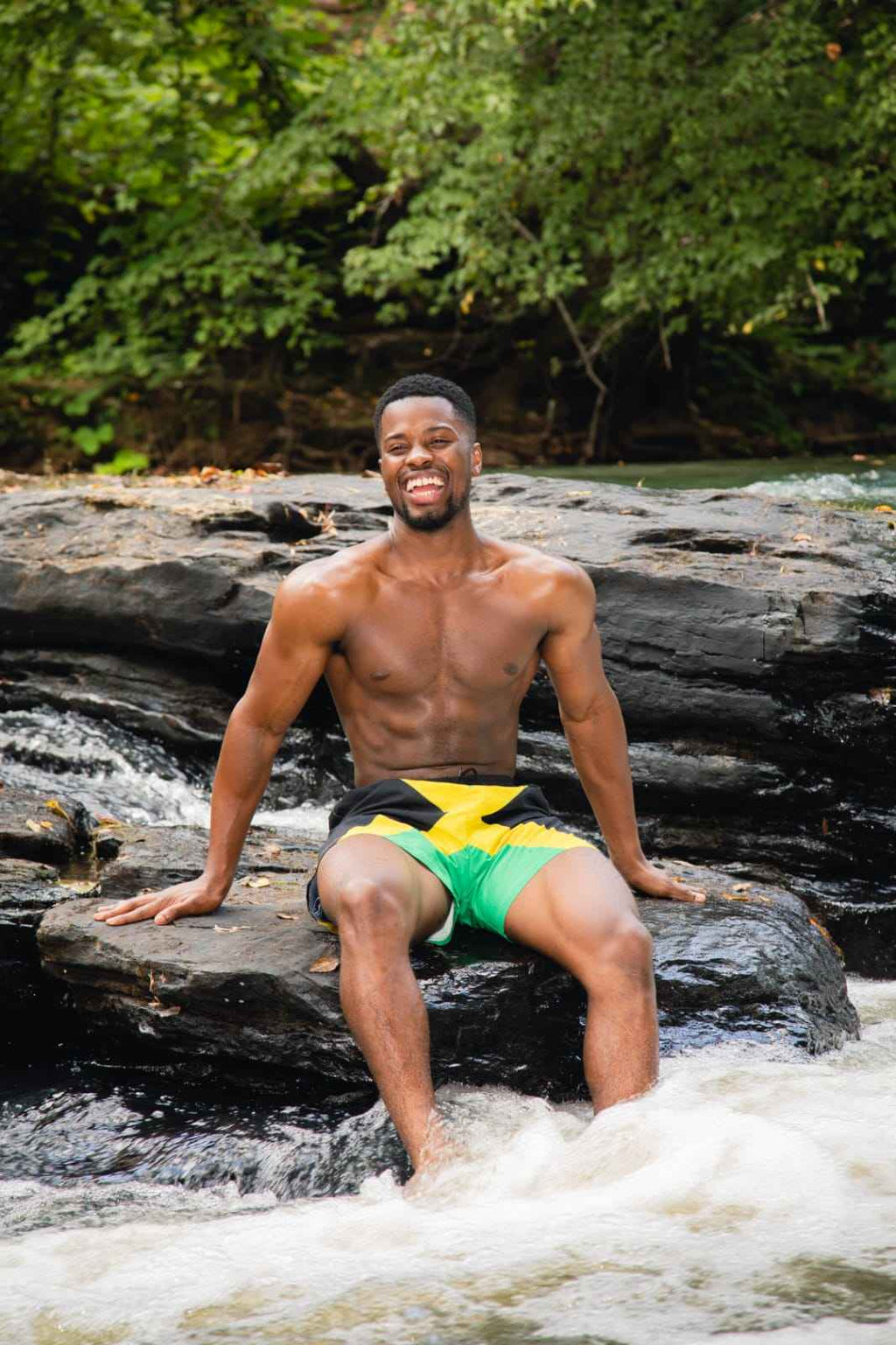 Smiling male model in black, green, and yellow Travaly Jamaica swim shorts on light background.