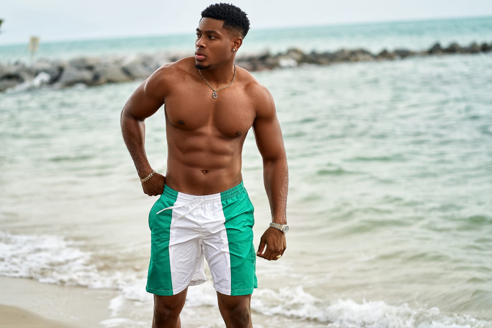 Model Ceasar Chukwuma wearing Nigegria swim shorts standing on a beach with ocean in the background
